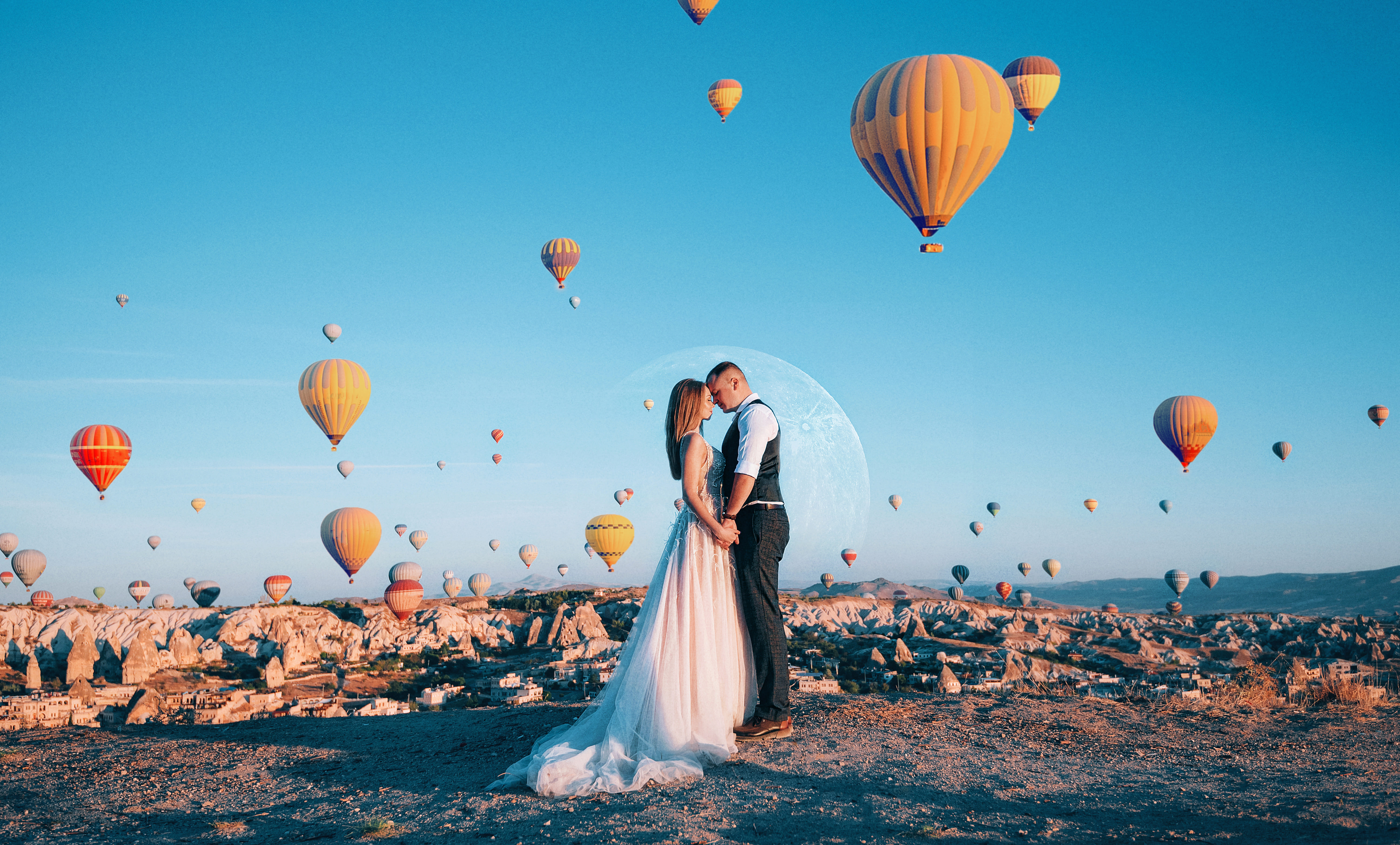 “A smiling bride and groom standing outdoors in Cappadocia with vibrant hot air balloons floating in the sky behind them.”