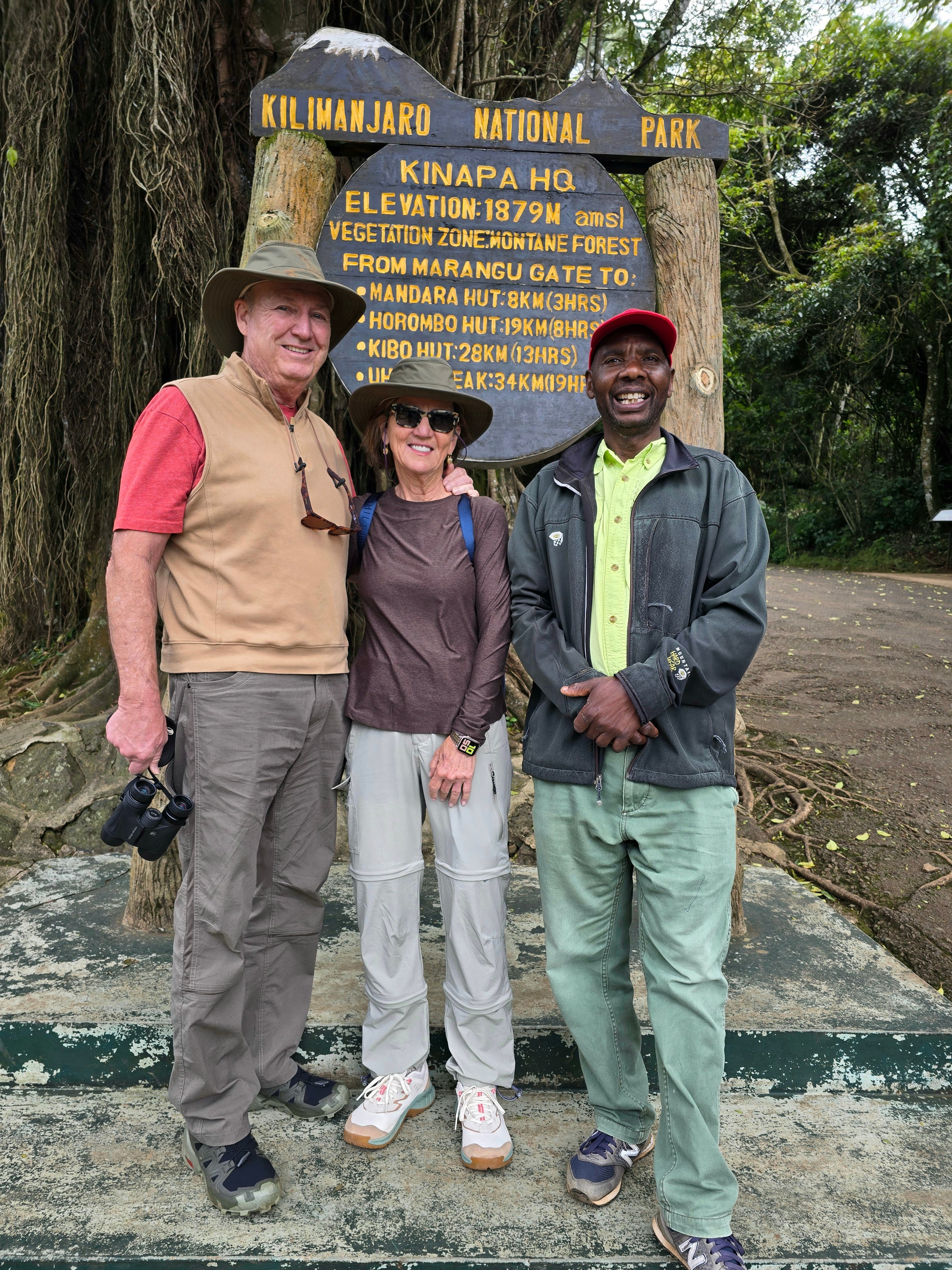A mountain guide standing together with two smiling tourists near the entrance gate sign for a Mount Kilimanjaro trek.