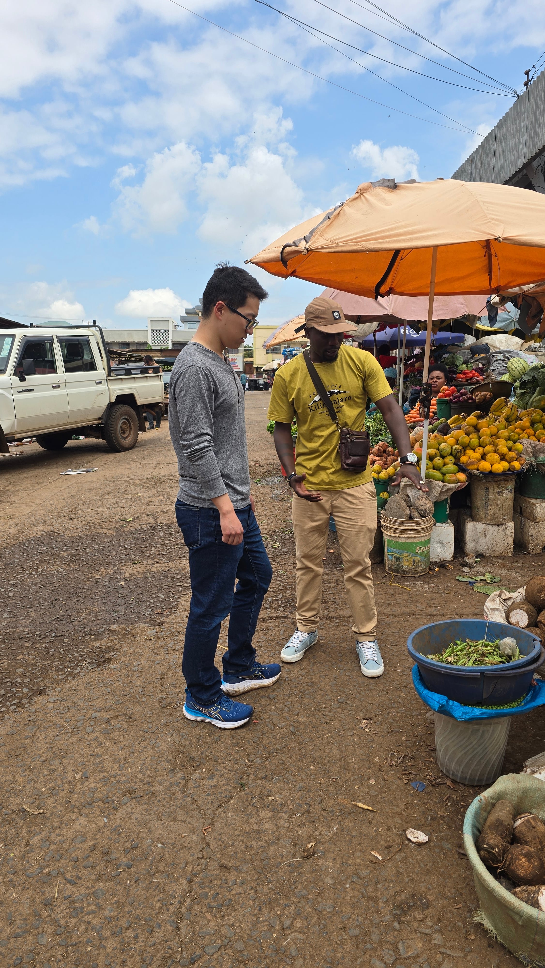 A guide showing a traveler around an outdoor fruit stall in Moshi town market.
