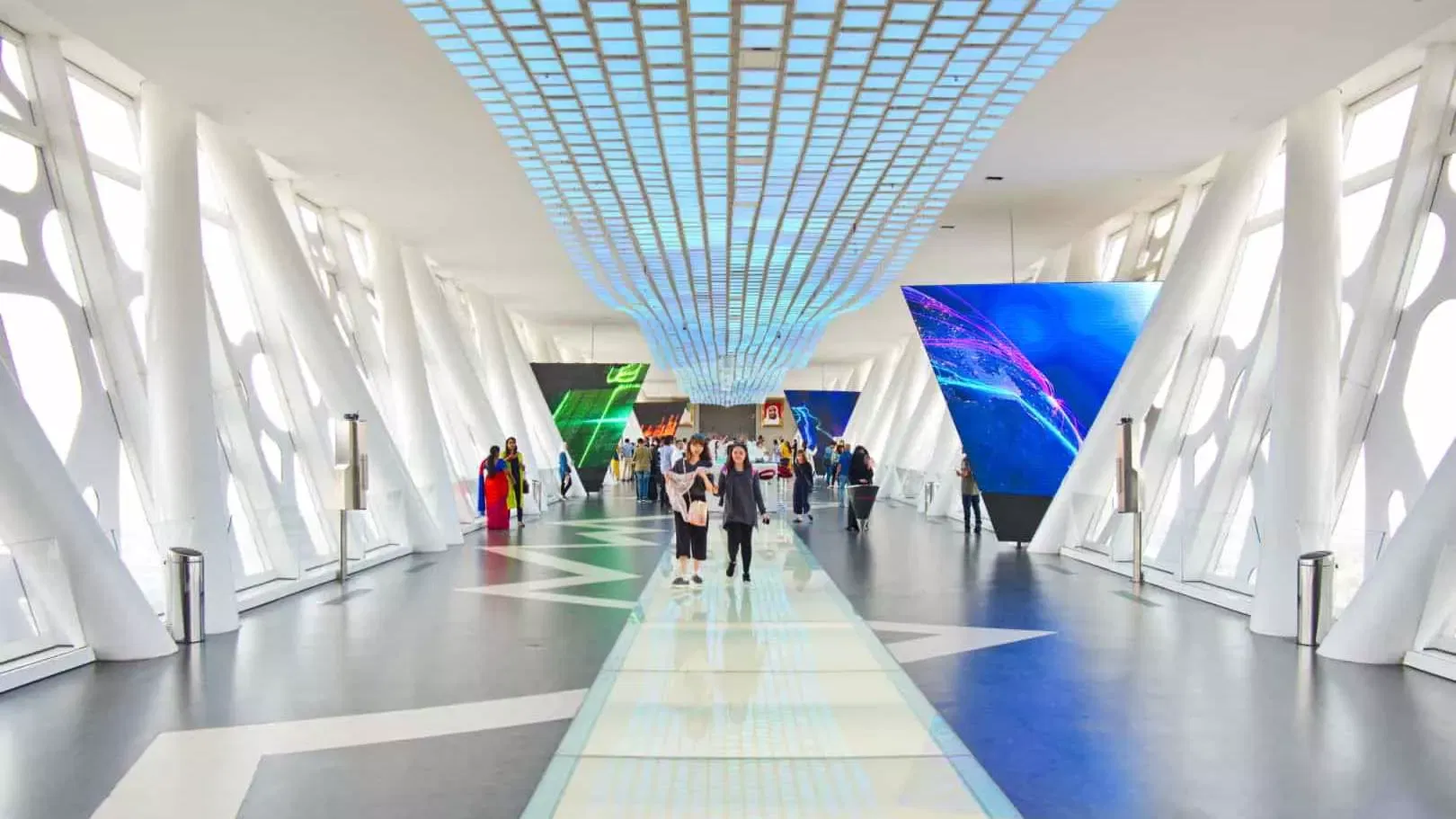 Visitors walking the bright white sky deck corridor along a glass-panelled walkway, with illuminated triangular screens on either side at Dubai Frame