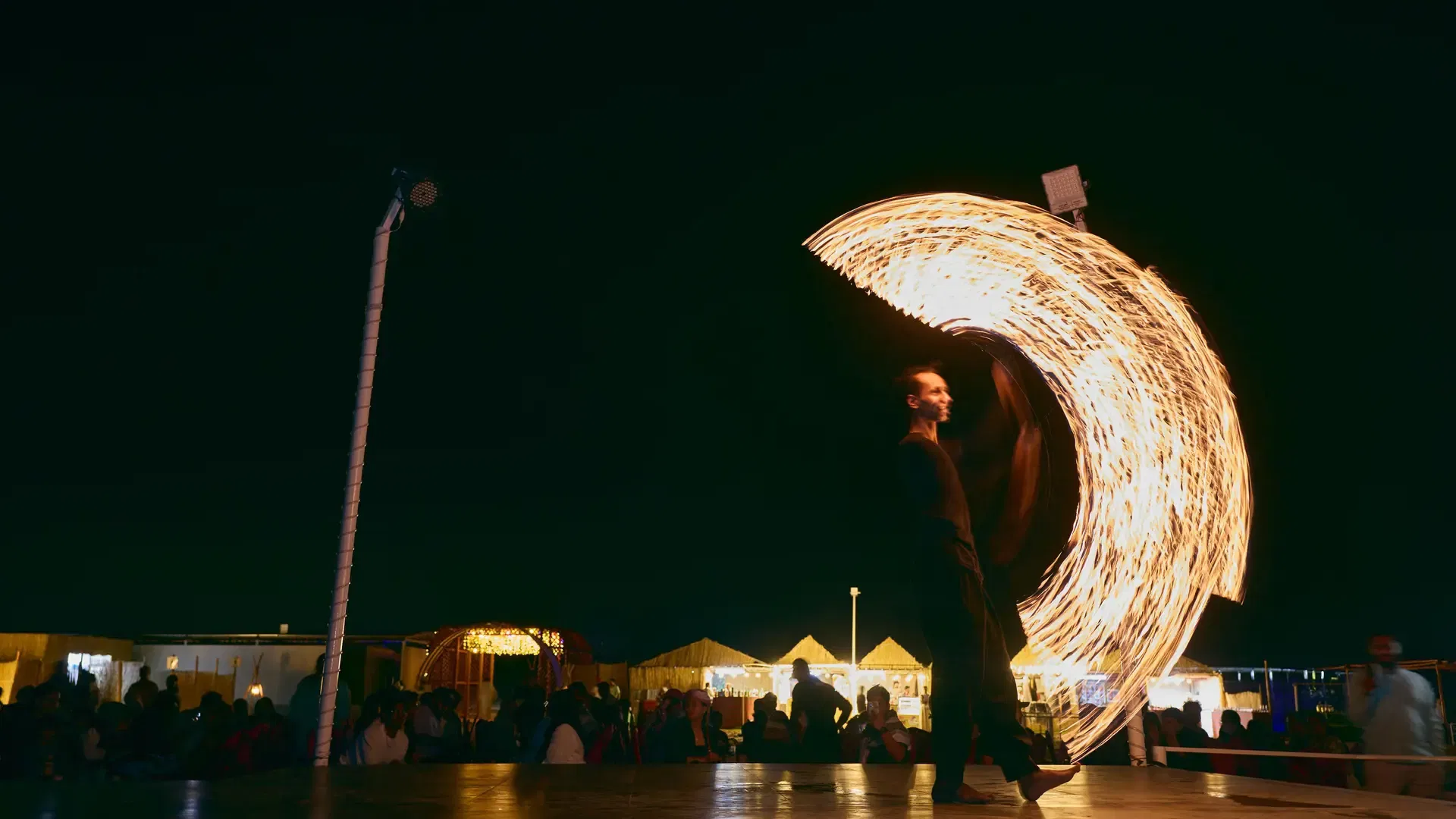 A male fire performer creates a spinning crescent of fire with a lit prop on stage at night at Desert Safari + Al Faqaa Camp.