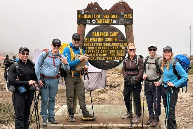 A group of six hikers posing with trekking poles in front of the Barranco Camp wooden sign in Kilimanjaro National Park.