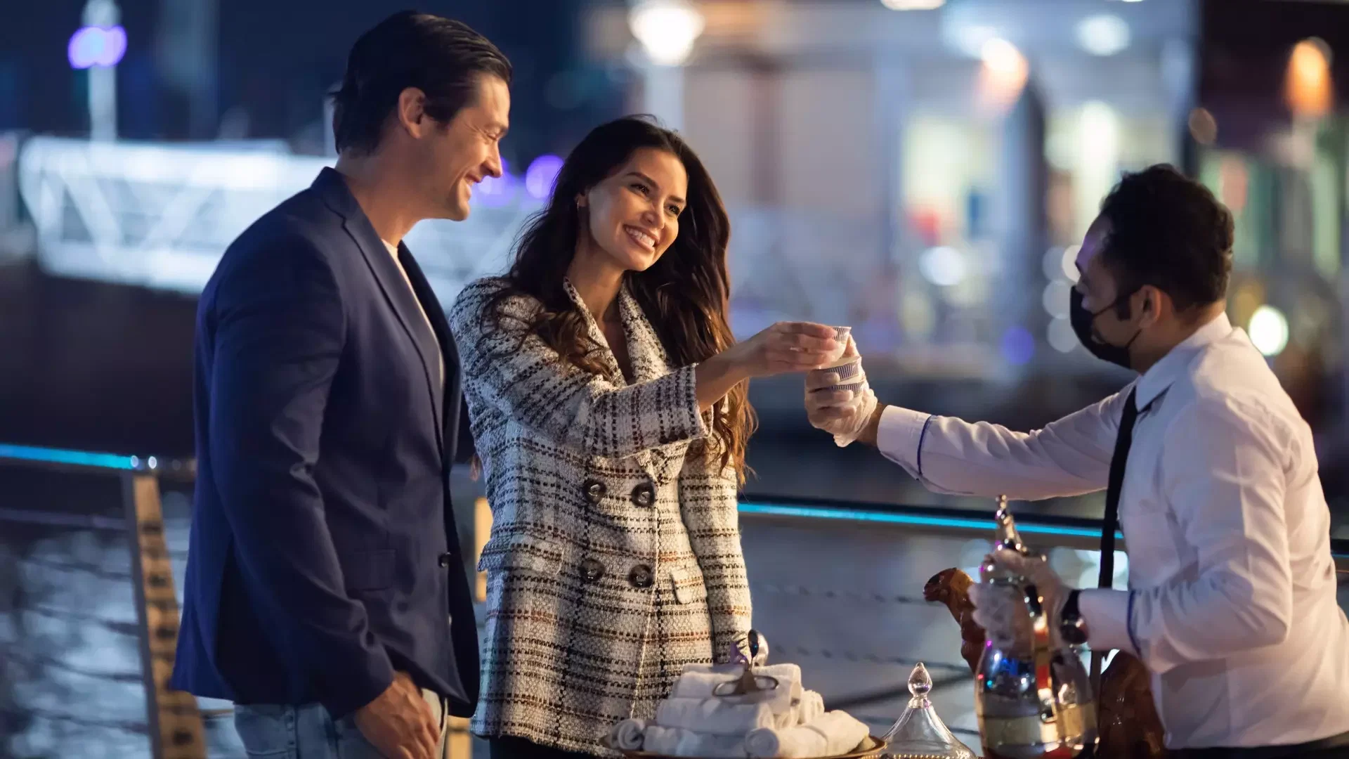 A man and a woman smile as they are served drinks by a staff member on the deck at Dubai Water Canal Dhow Cruise.