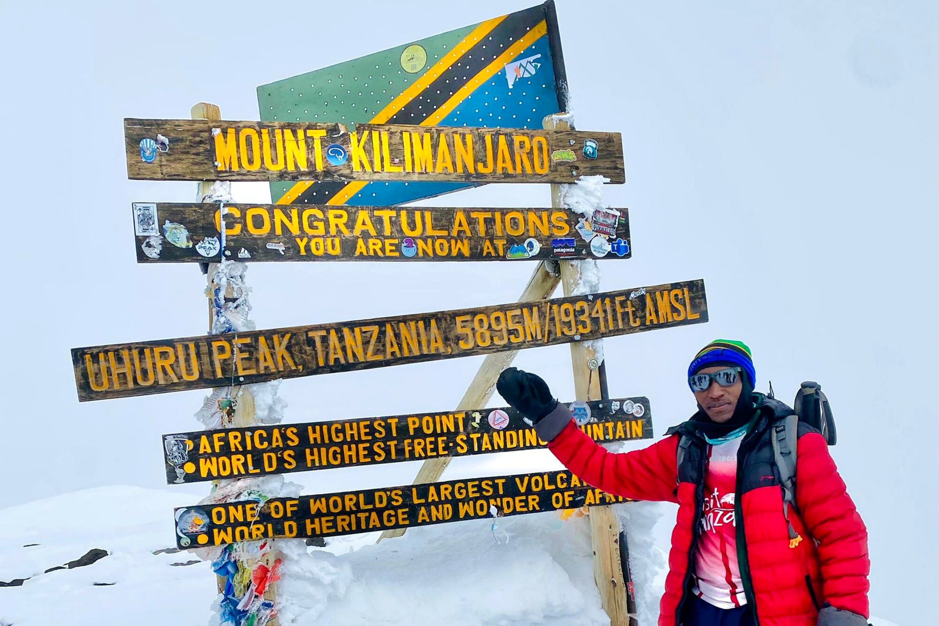 A hiker in a red jacket standing next to the wooden Mount Kilimanjaro summit sign at Uhuru Peak surrounded by snow.