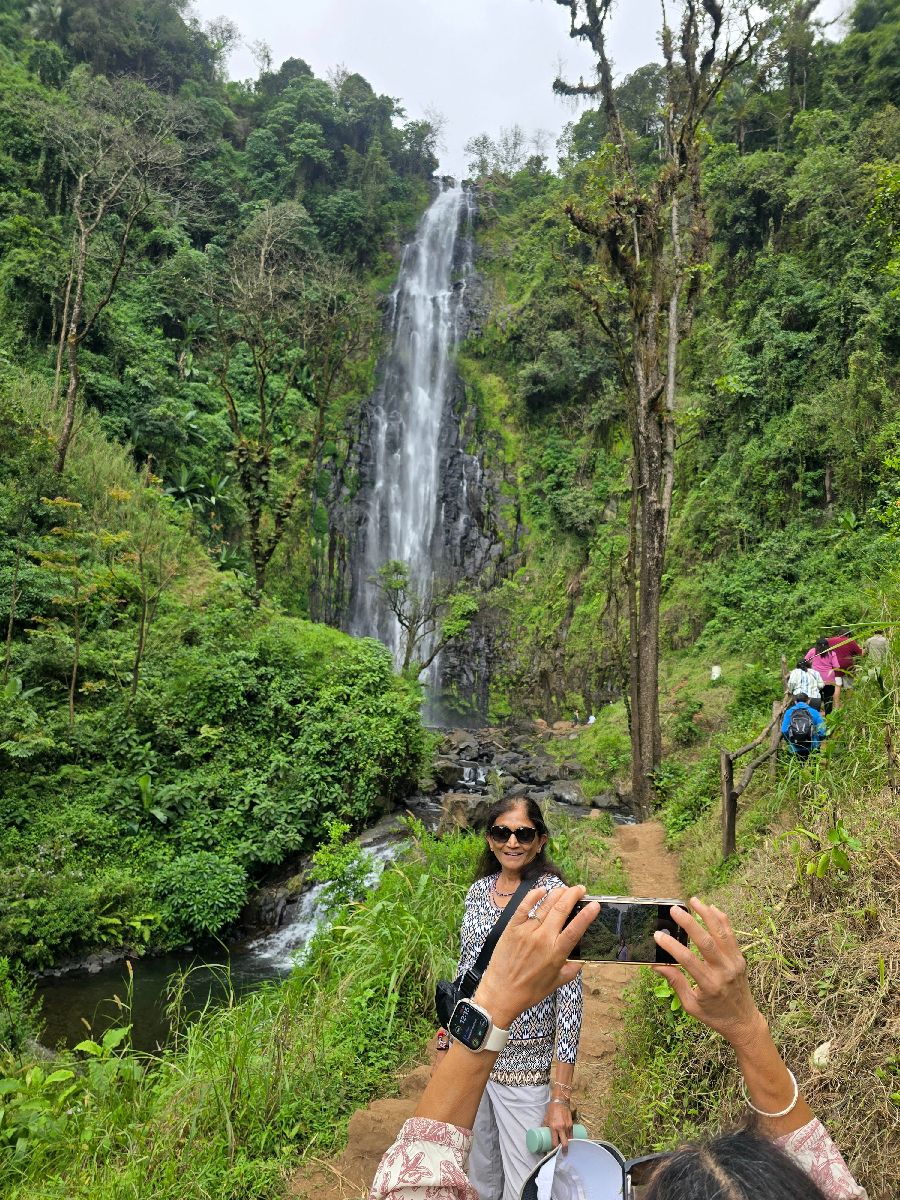 Traveler photographing the towering Materuni Waterfall surrounded by lush green tropical foliage.