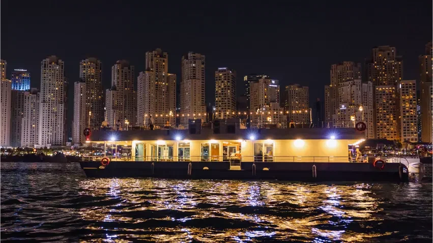 A large illuminated boat cruises on water with its reflection shimmering beneath a backdrop of tall skyscrapers at Alexandra Sea Lounge.