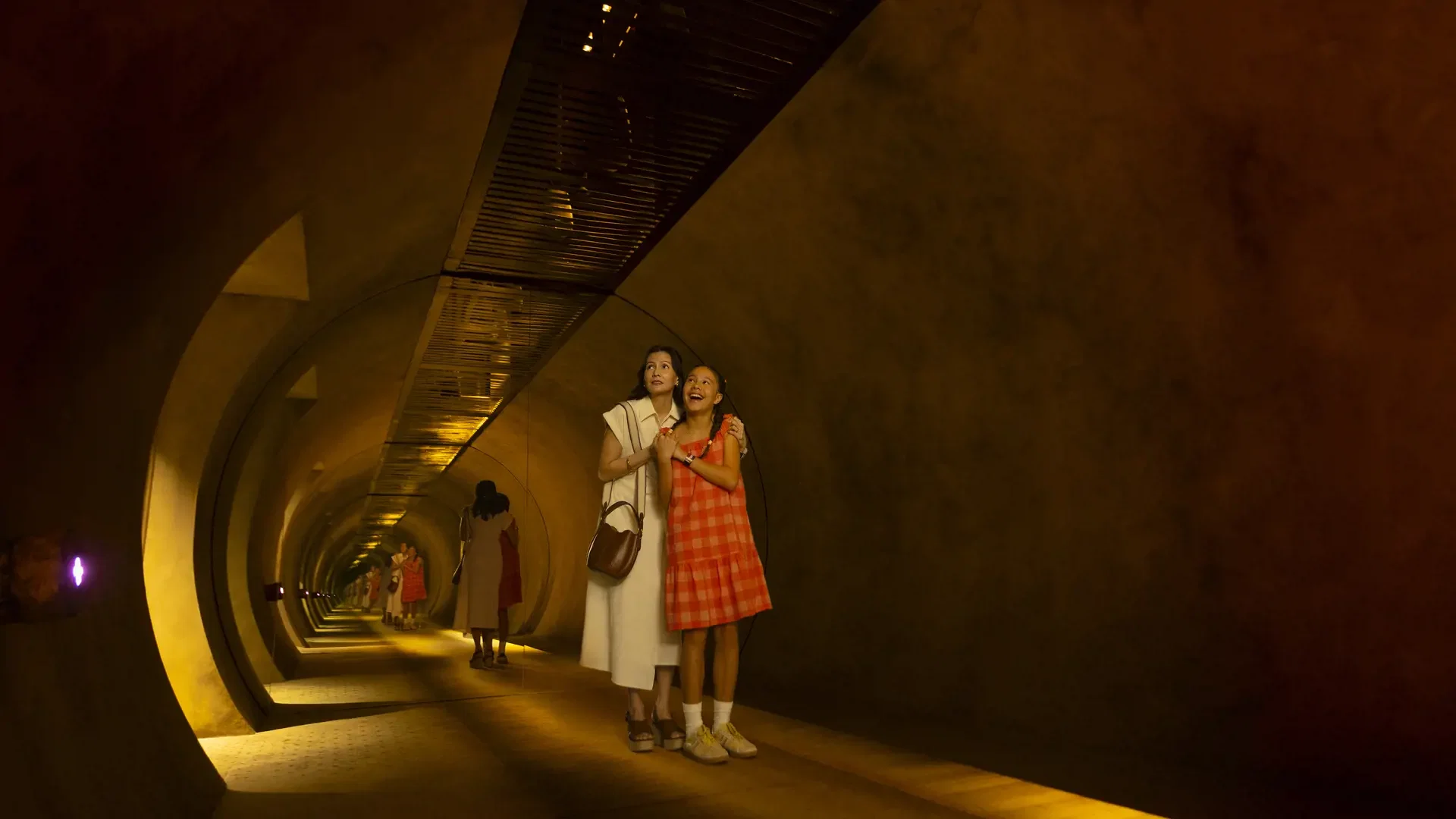 A woman and a young girl stand arm-in-arm inside a curved, dimly lit tunnel with arched ceilings at Exploria in Mandai Singapore Ticket.