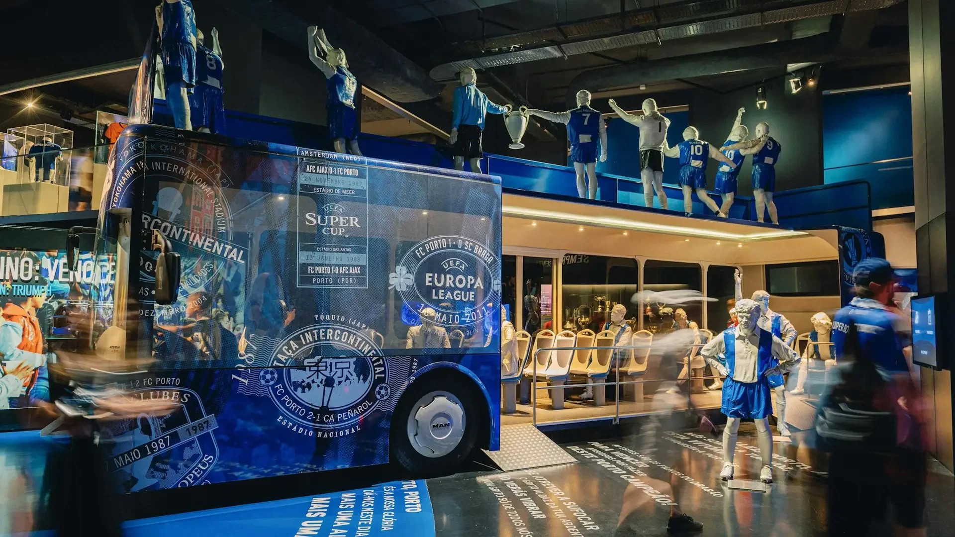 A large decorated blue bus display features several mannequin figures standing on its roof inside a brightly lit exhibition hall at FC Porto Museum & Dragão Stadium ticket.