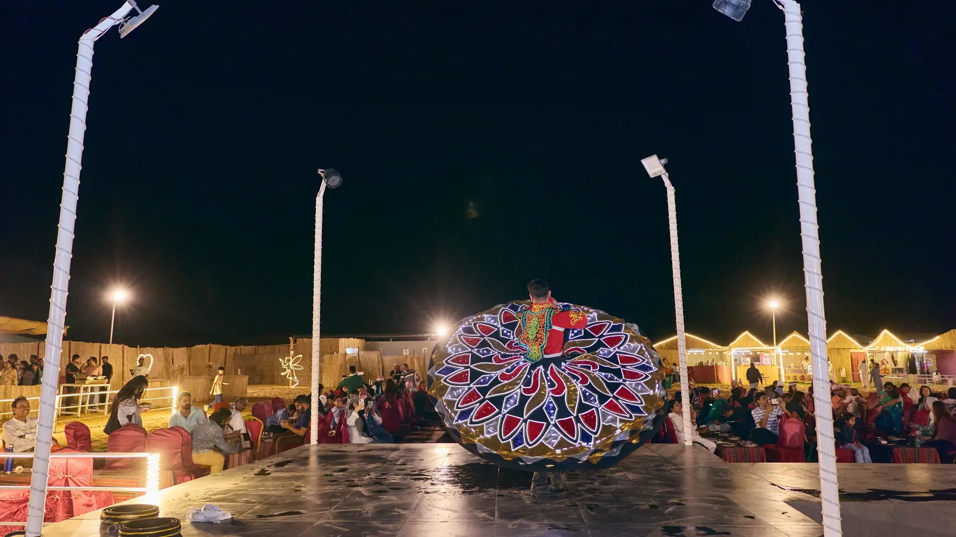 A male dancer performs a spinning routine in a brightly decorated costume on a lit stage at Desert Safari + Al Faqaa Camp.