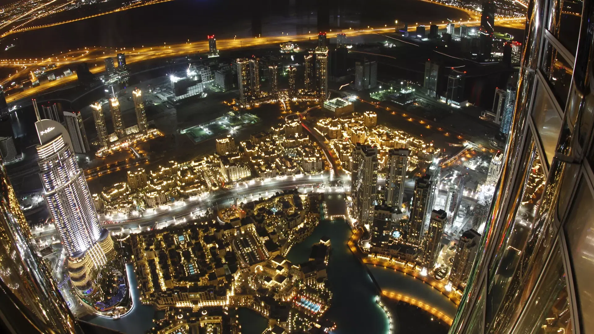 Wide-angle night view of Downtown Dubai seen past the curved glass and steel of the tower's own facade at Burj Khalifa