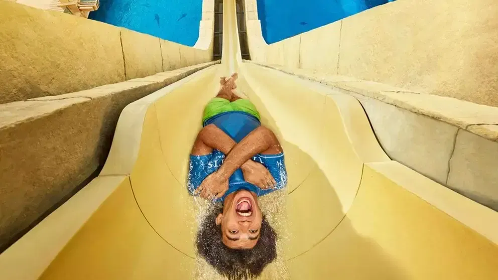 A rider in blue and green swim shorts lying back and laughing as she rushes down a wide sand-coloured slide chute at Aquaventure Waterpark