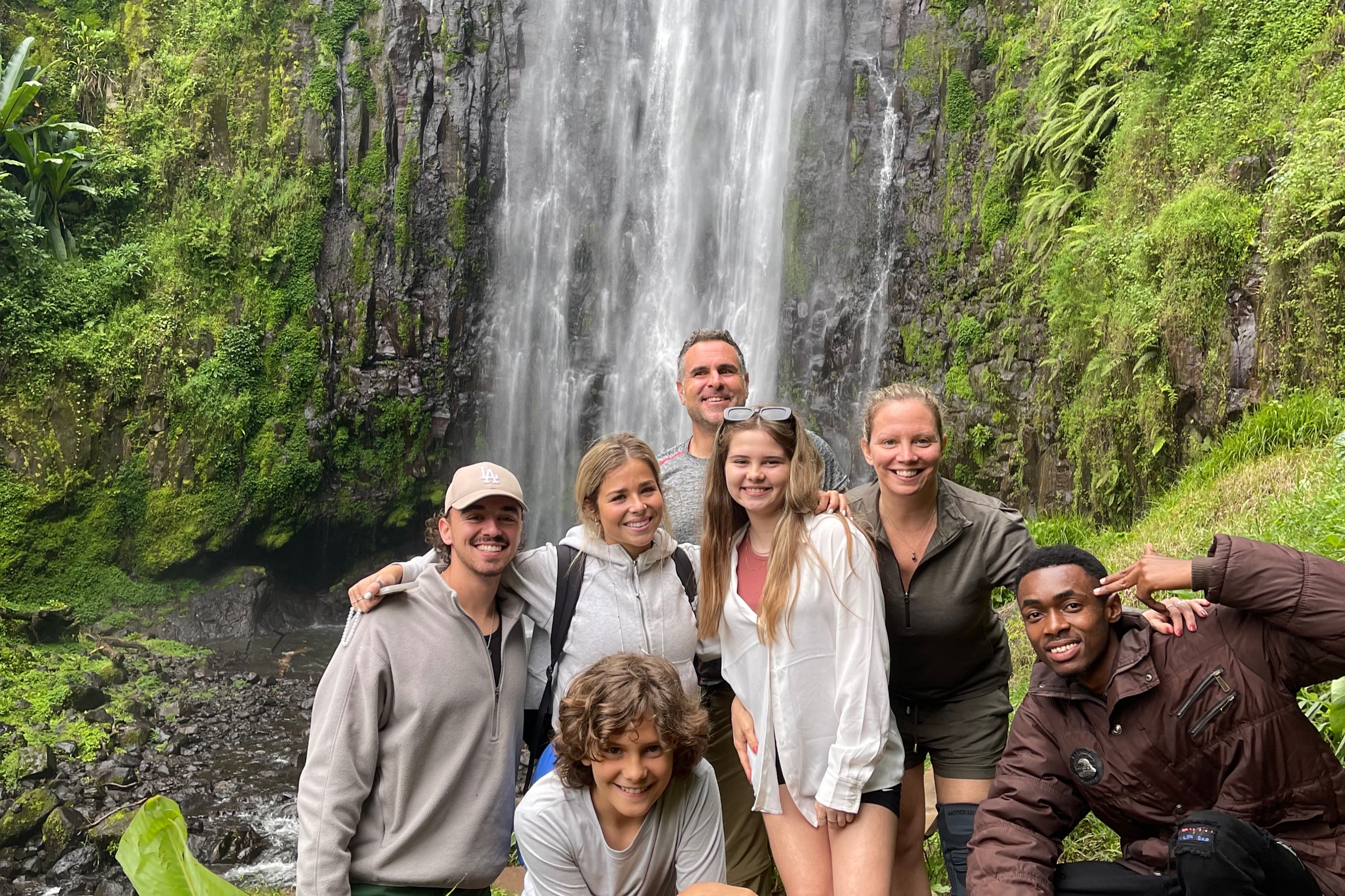 A smiling group of multi-ethnic visitors posing together in front of Materuni Waterfalls near Moshi.