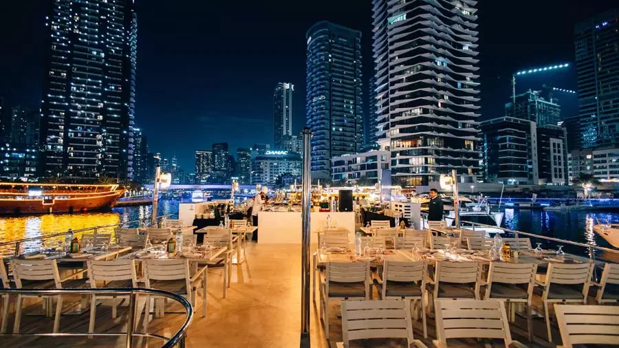 On an open-air deck at night, multiple tables are arranged with white chairs under the glow of overhead lights, against a backdrop of illuminated high-rise buildings at Alexandra Sea Lounge.