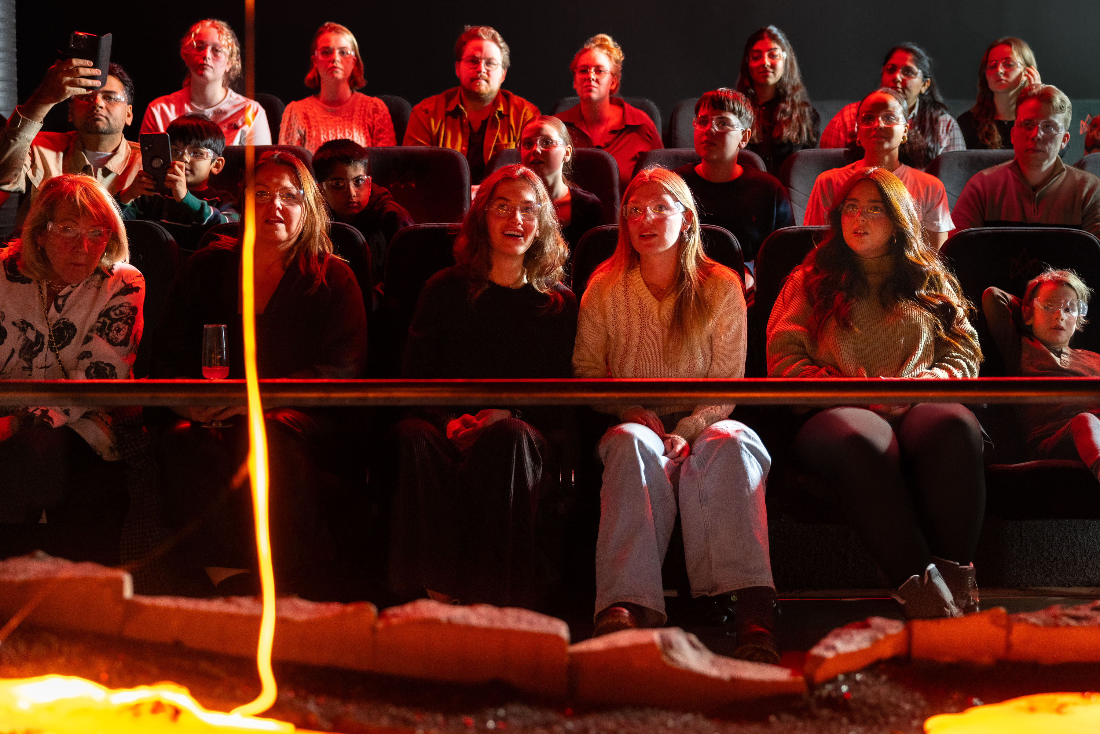 Audience members watch closely and take photos as a large, glowing blob of cooled lava rests on a sand platform in front of them.