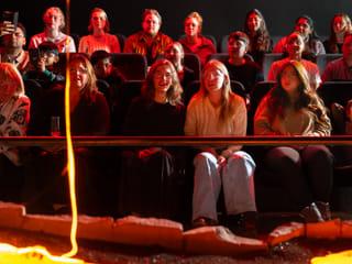 Audience members watch closely and take photos as a large, glowing blob of cooled lava rests on a sand platform in front of them.