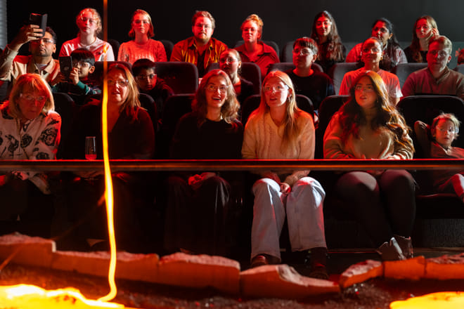 Audience members watch closely and take photos as a large, glowing blob of cooled lava rests on a sand platform in front of them.