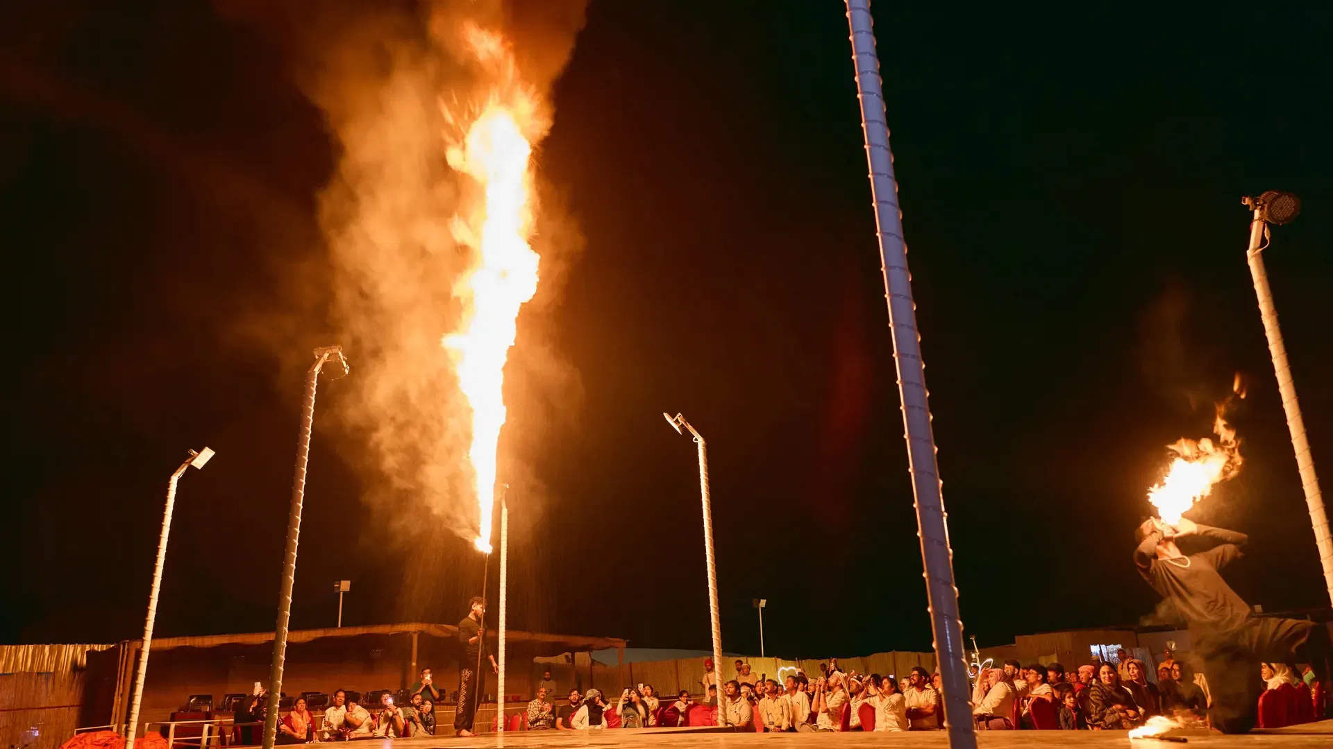 A performer on a stage breathes a plume of fire into the night air while an audience watches at Desert Safari + Al Faqaa Camp.