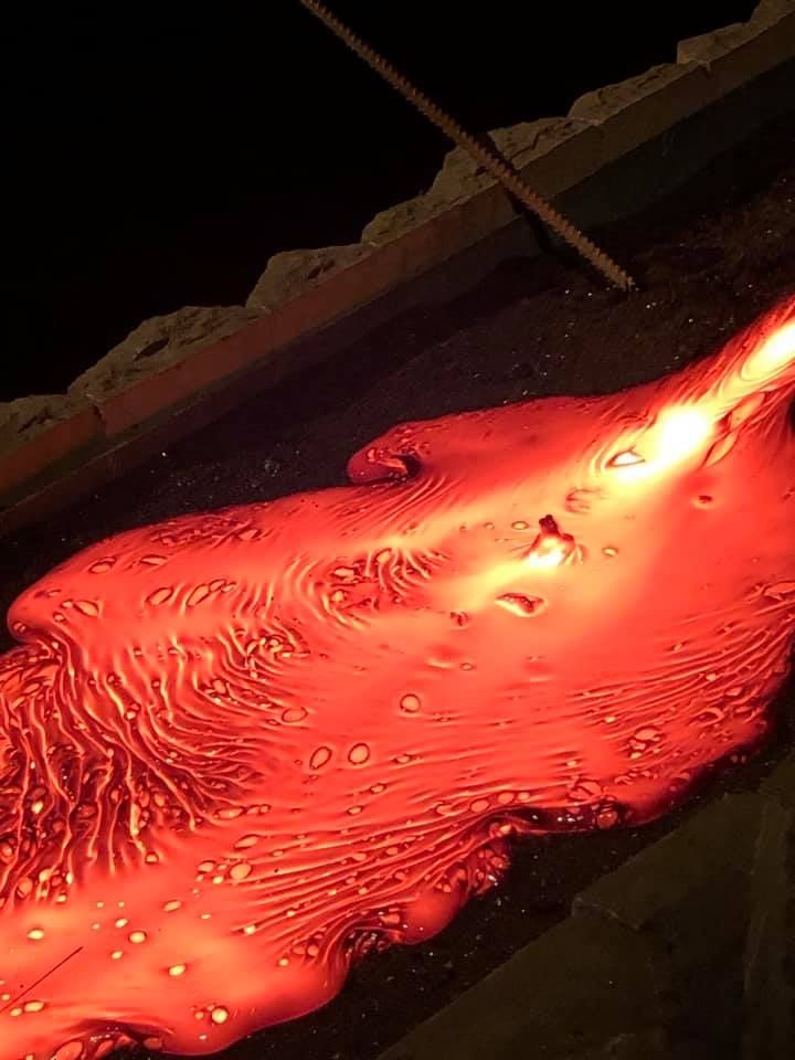 A close-up image of molten lava flowing across a dark sandbed, showing glowing orange textures, swirling lines, and bubbles as the lava spre