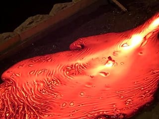 A close-up image of molten lava flowing across a dark sandbed, showing glowing orange textures, swirling lines, and bubbles as the lava spre