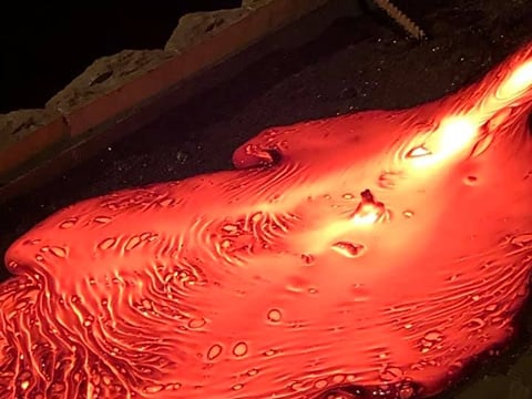 A close-up image of molten lava flowing across a dark sandbed, showing glowing orange textures, swirling lines, and bubbles as the lava spre