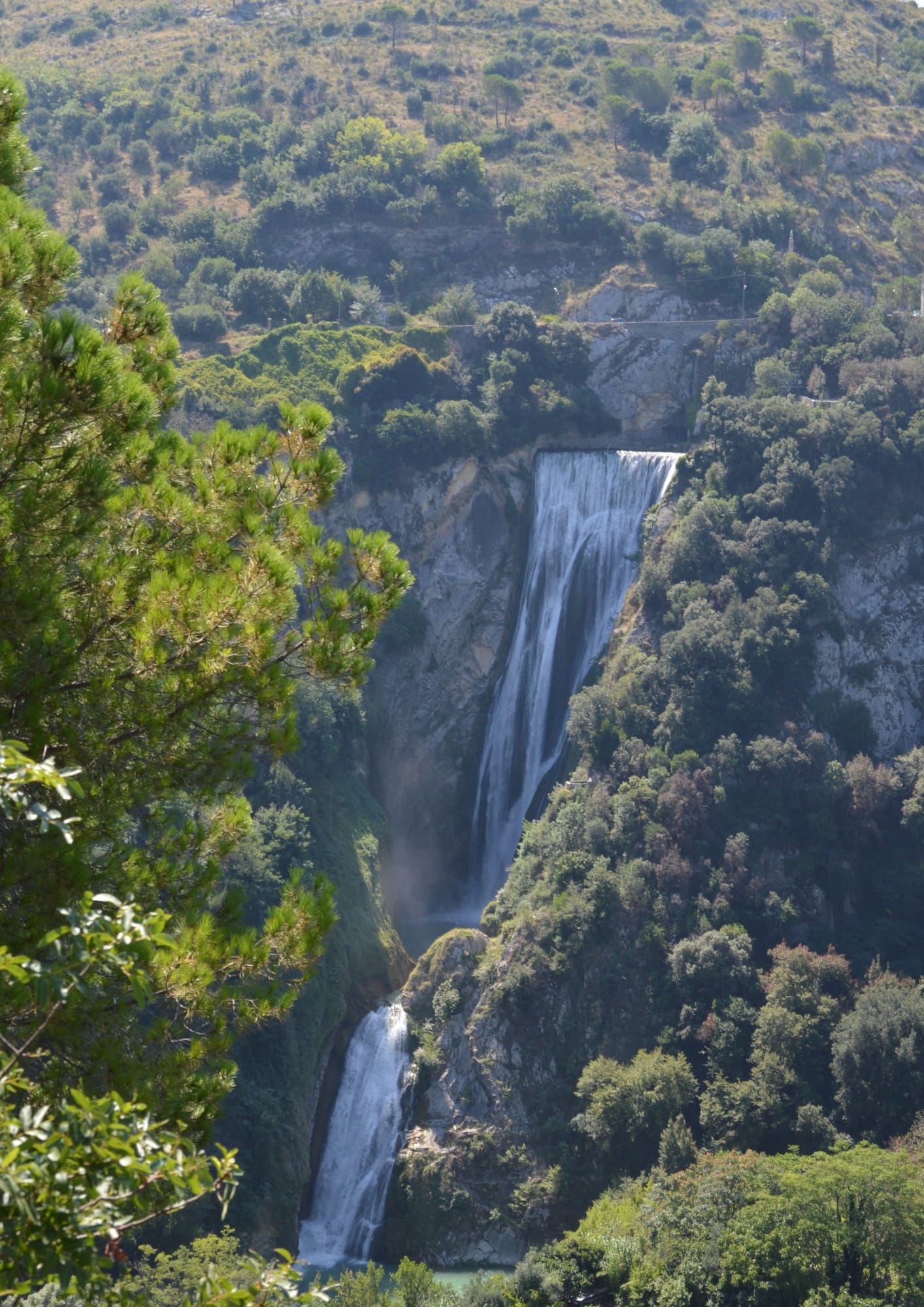 Distant view of a tall waterfall dropping through a wooded gorge, with hillside buildings in the distance.