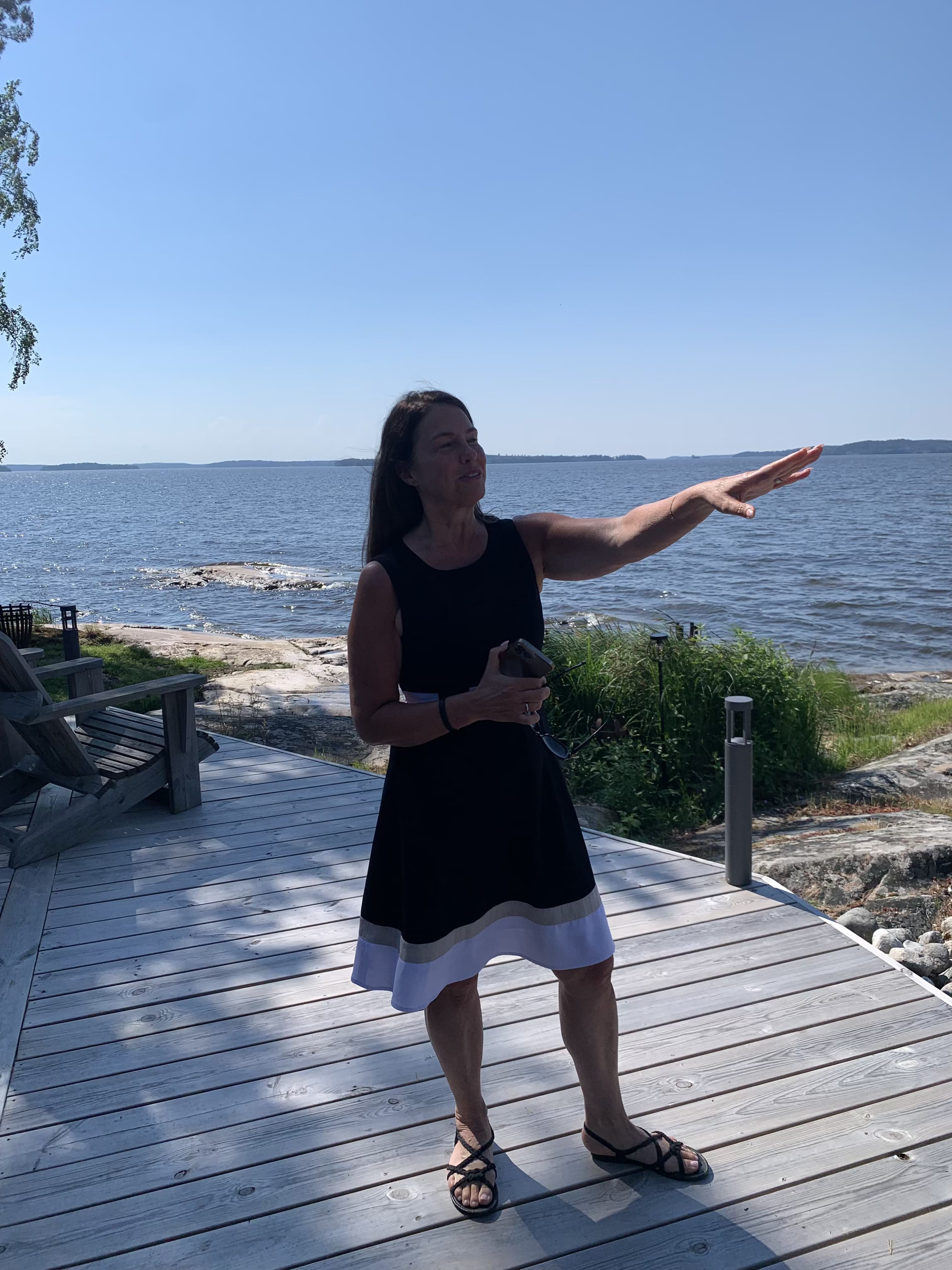 A woman in a sleeveless black dress with white trim stands on a wooden deck, gesturing toward the water on a sunny day.