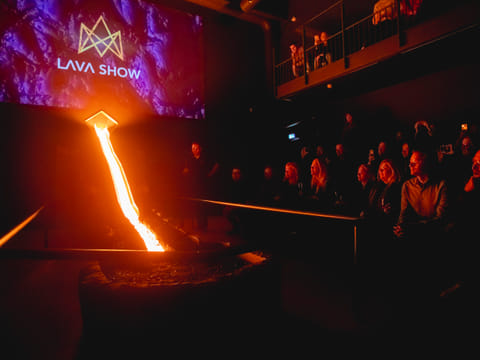 Molten lava flows down a steel channel in a dark showroom with the "Lava Show" logo projected on a screen, while the audience watches from b