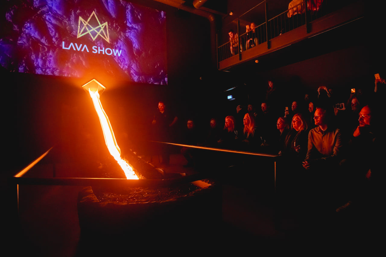 Molten lava flows down a steel channel in a dark showroom with the "Lava Show" logo projected on a screen, while the audience watches from b