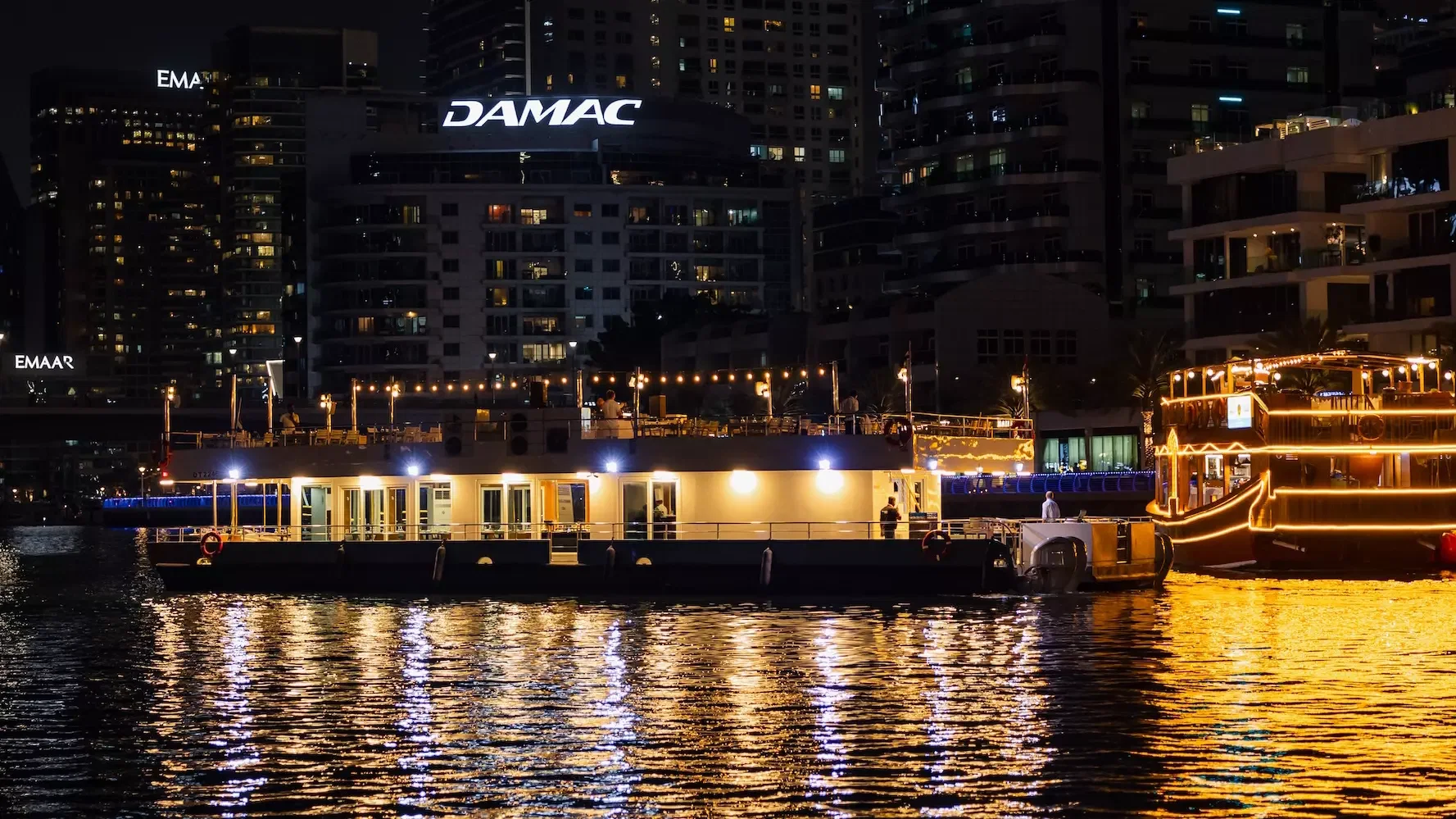 A brightly lit floating restaurant is docked on the water at night with city buildings in the background at Alexandra Sea Lounge.