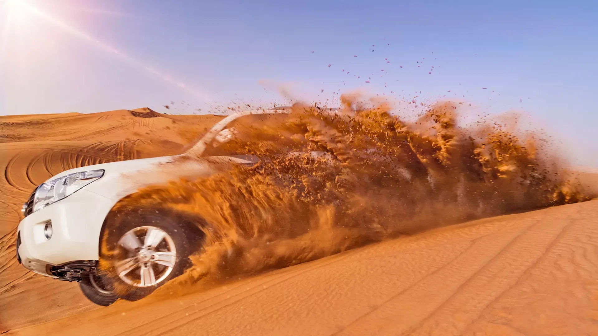 A white SUV drifts across a sandy slope, sending a thick plume of sand billowing into the air against a blue sky at Desert Safari + Al Faqaa Camp.