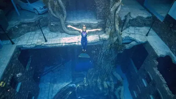 A scuba diver floats vertically with arms outstretched inside an elaborate underwater sunken city featuring a submerged car and massive tree at Deep Dive Dubai.