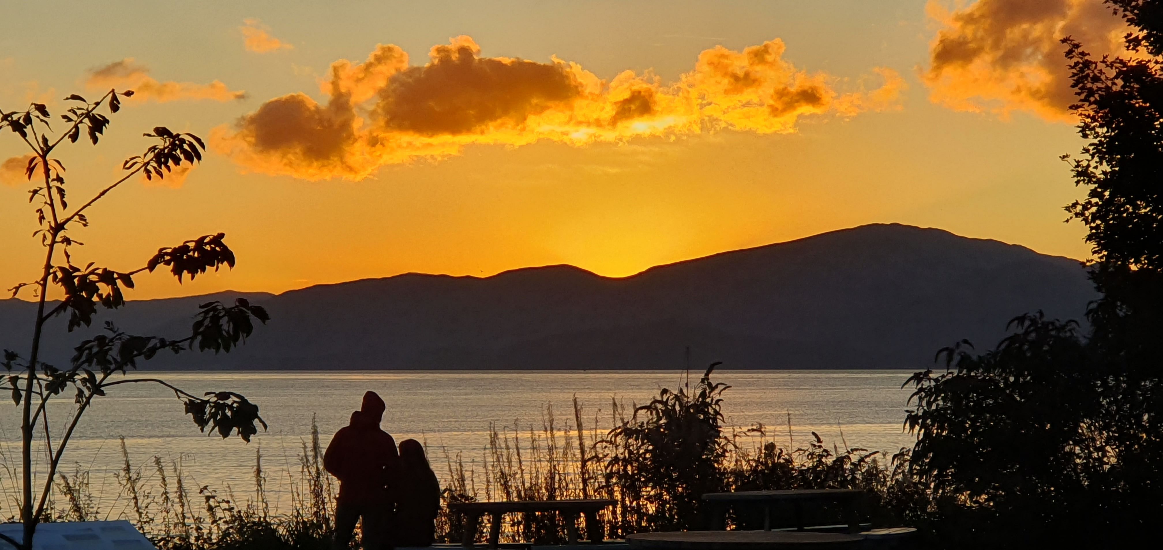 Couple watching the sunset from the shore of a loch in Scotland. The golden sky has clouds and the horizon is silhouetted beautifully.