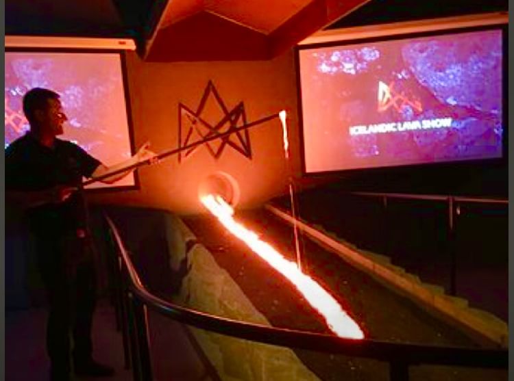A presenter stands beside a glowing stream of molten lava flowing from a furnace into a trench, with screens displaying