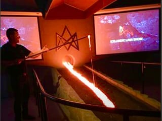 A presenter stands beside a glowing stream of molten lava flowing from a furnace into a trench, with screens displaying "Icelandic Lava Show