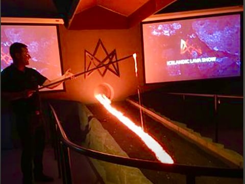 A presenter stands beside a glowing stream of molten lava flowing from a furnace into a trench, with screens displaying "Icelandic Lava Show