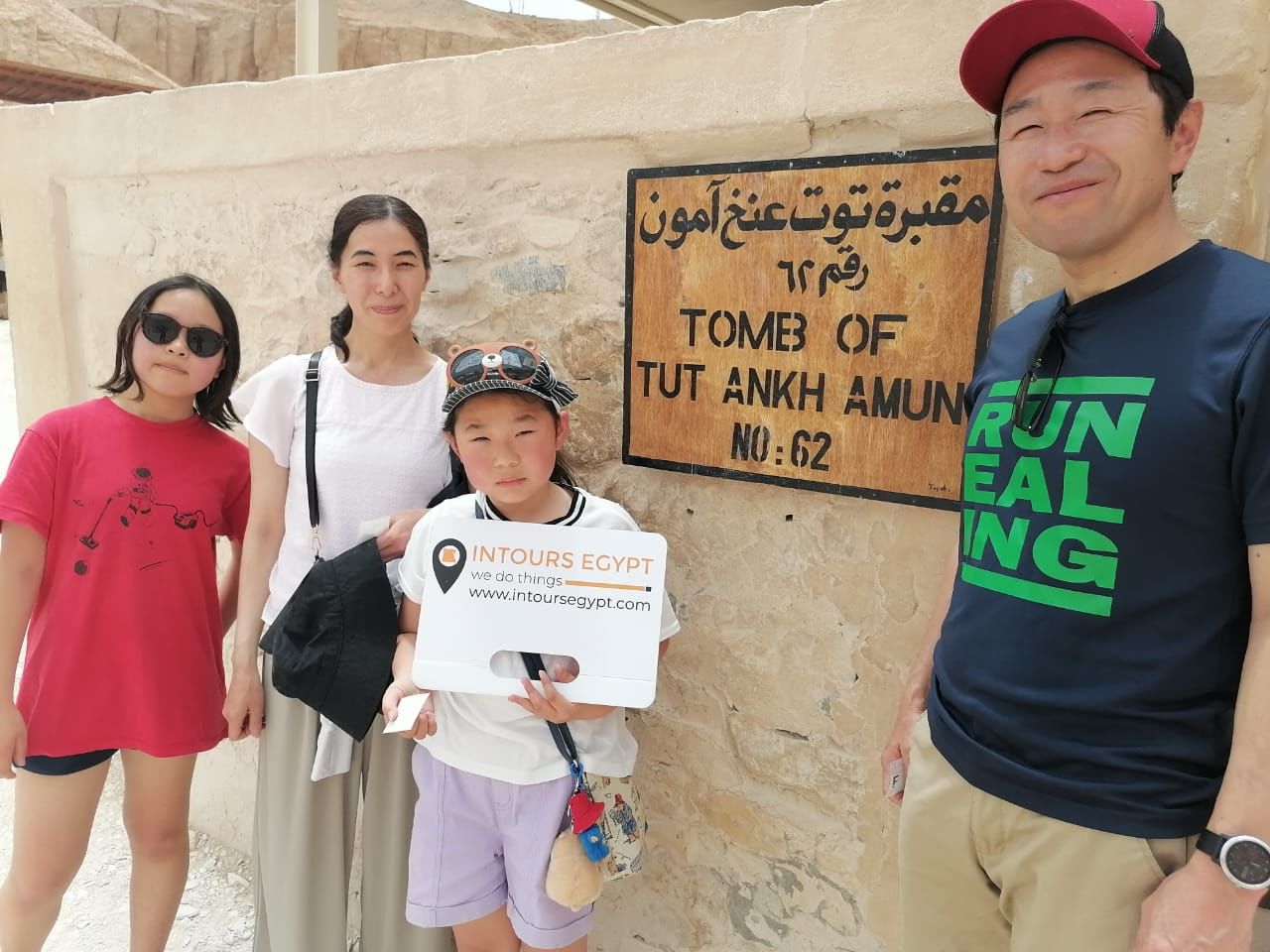 Une famille de quatre personnes se tient debout, souriante, à côté du panneau indiquant la tombe de Toutankhamon, dans la Vallée des Rois, avec un enfant tenant un Intours E