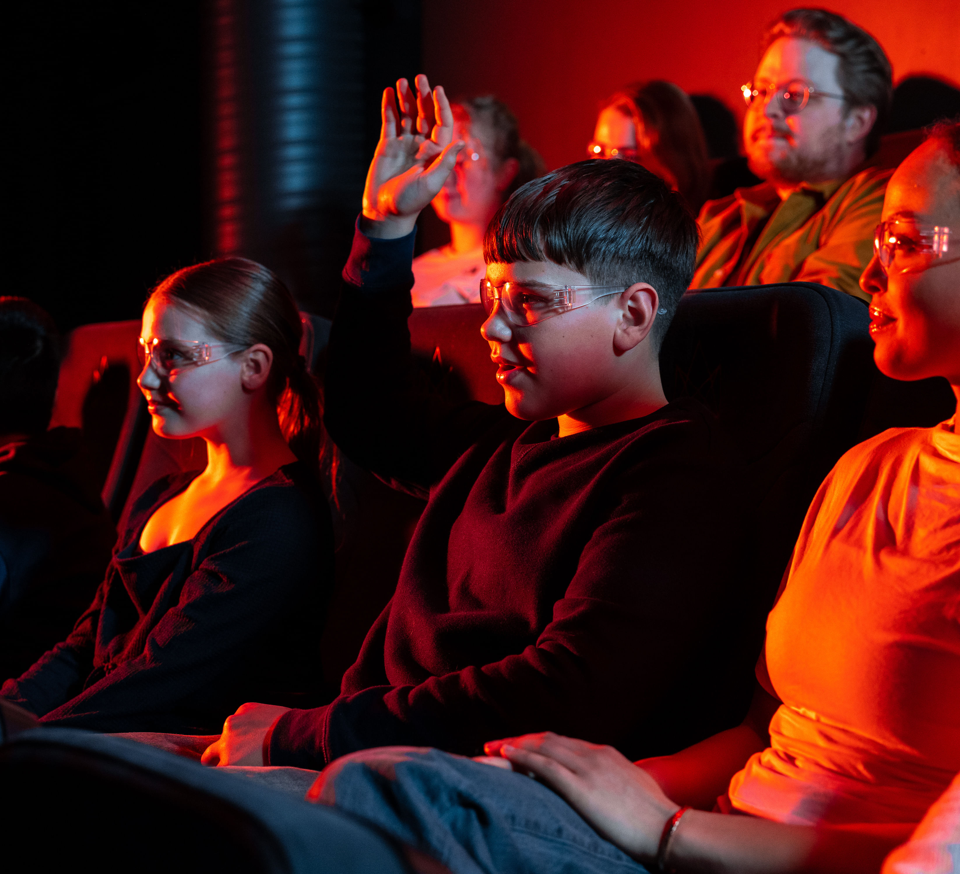 A young boy raises his hand during the Lava Show while sitting in a dark theater with other children and adults, all wearing safety glasses