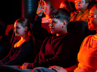 A young boy raises his hand during the Lava Show while sitting in a dark theater with other children and adults, all wearing safety glasses