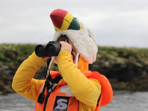 A child in a puffin hat and life vest watches puffins through binoculars.