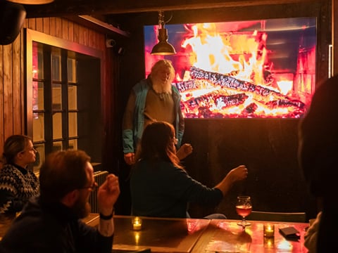 A man with long white hair giving a presentation in front of a TV displaying a cozy fireplace scene
