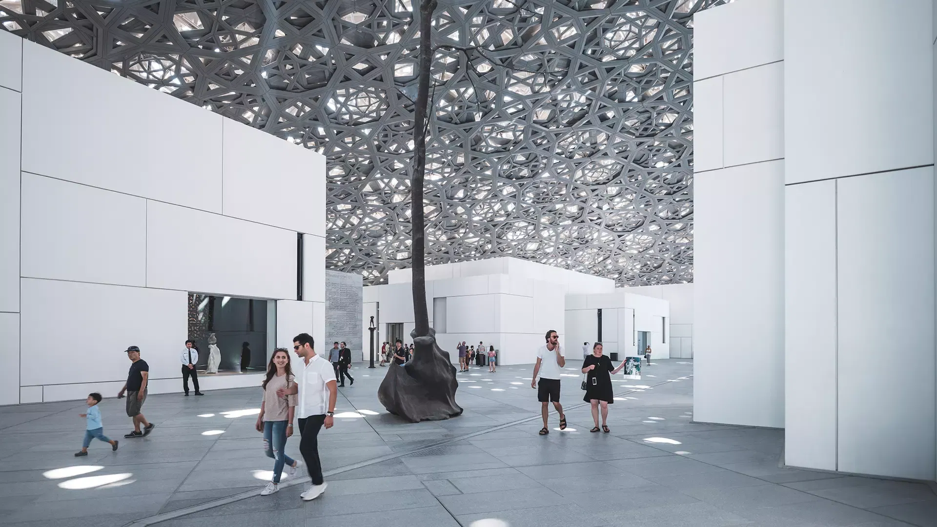 A couple walking past a tall bronze tree sculpture standing in the open museum square beneath the star-patterned canopy at Louvre Abu Dhabi