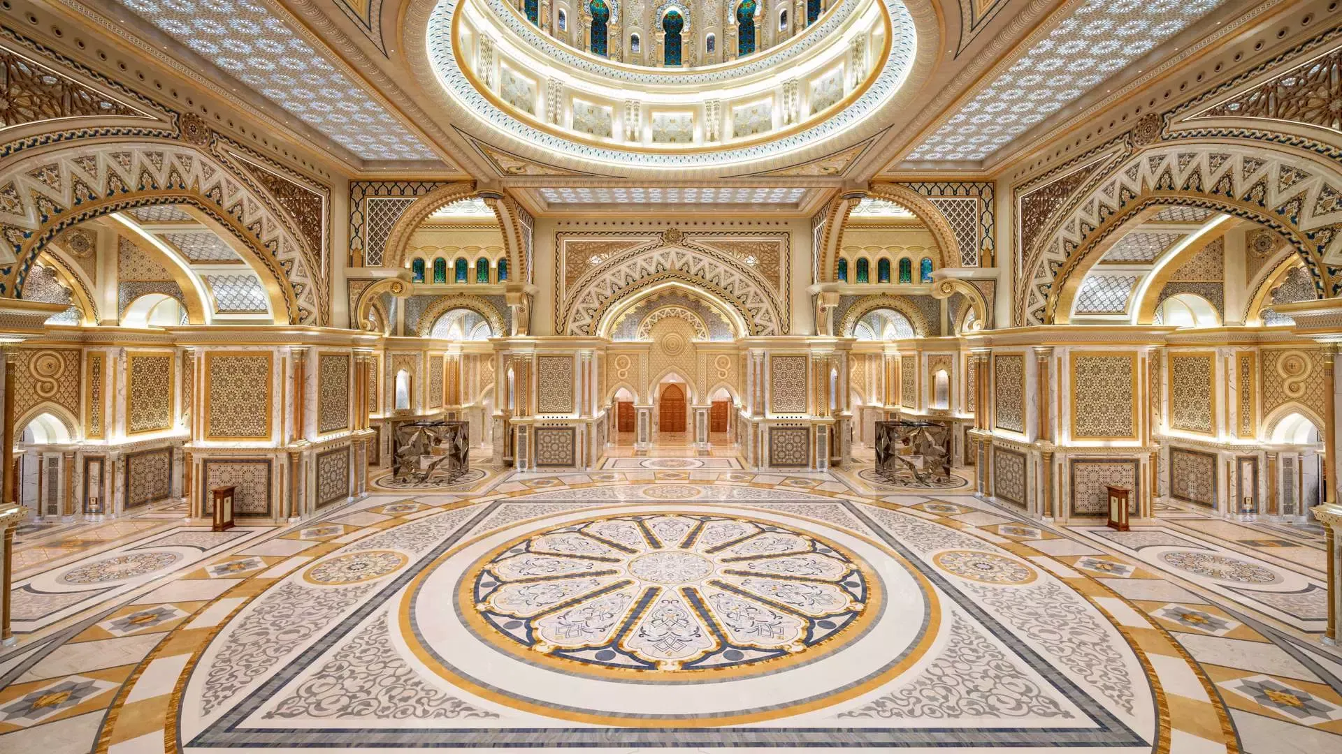A grand, symmetrical interior hall featuring ornate white and gold archways, intricate geometric wall panels, and a large circular mosaic inlay on the polished marble floor at Qasr Al Watan, Royal Palace.