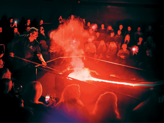 A Lava Show presenter uses a tool to manipulate a stream of glowing molten lava while a full audience watches under red light and rising ste