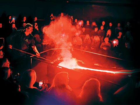 A Lava Show presenter uses a tool to manipulate a stream of glowing molten lava while a full audience watches under red light and rising ste