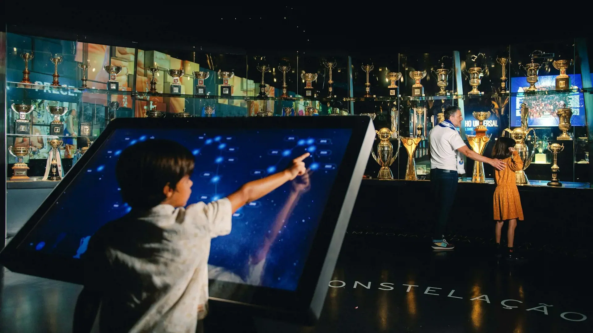 A young boy interacts with a large interactive touchscreen display while others look at trophy cases at FC Porto Museum & Dragão Stadium ticket.