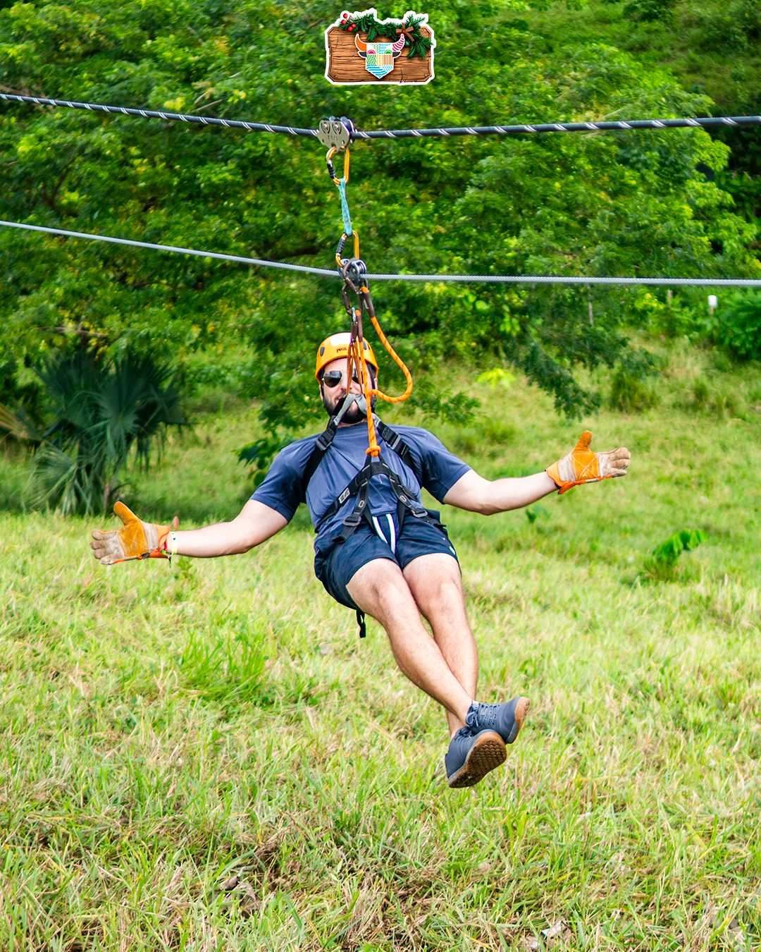 Man wearing a helmet, safety harness, and gloves gliding on a zip line over lush green scenery in Punta Cana, extending his arms and enjoyin