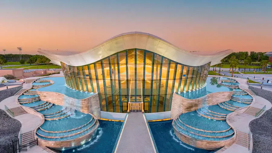 An exterior view showcases the glass-fronted building with a curved roof surrounded by tiered cascading pools at Deep Dive Dubai.
