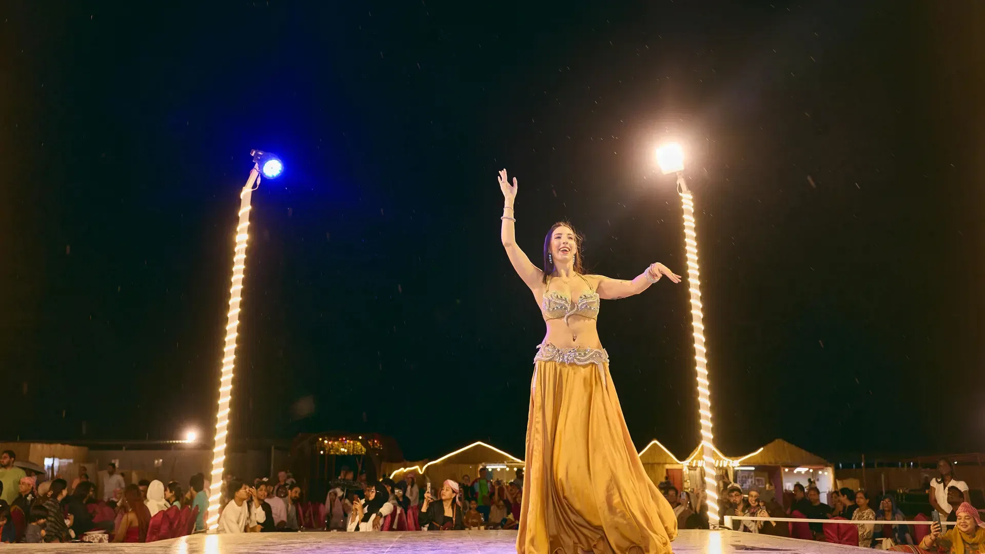 A woman in a gold belly-dancing costume performs on an outdoor stage at night at Desert Safari + Al Faqaa Camp.