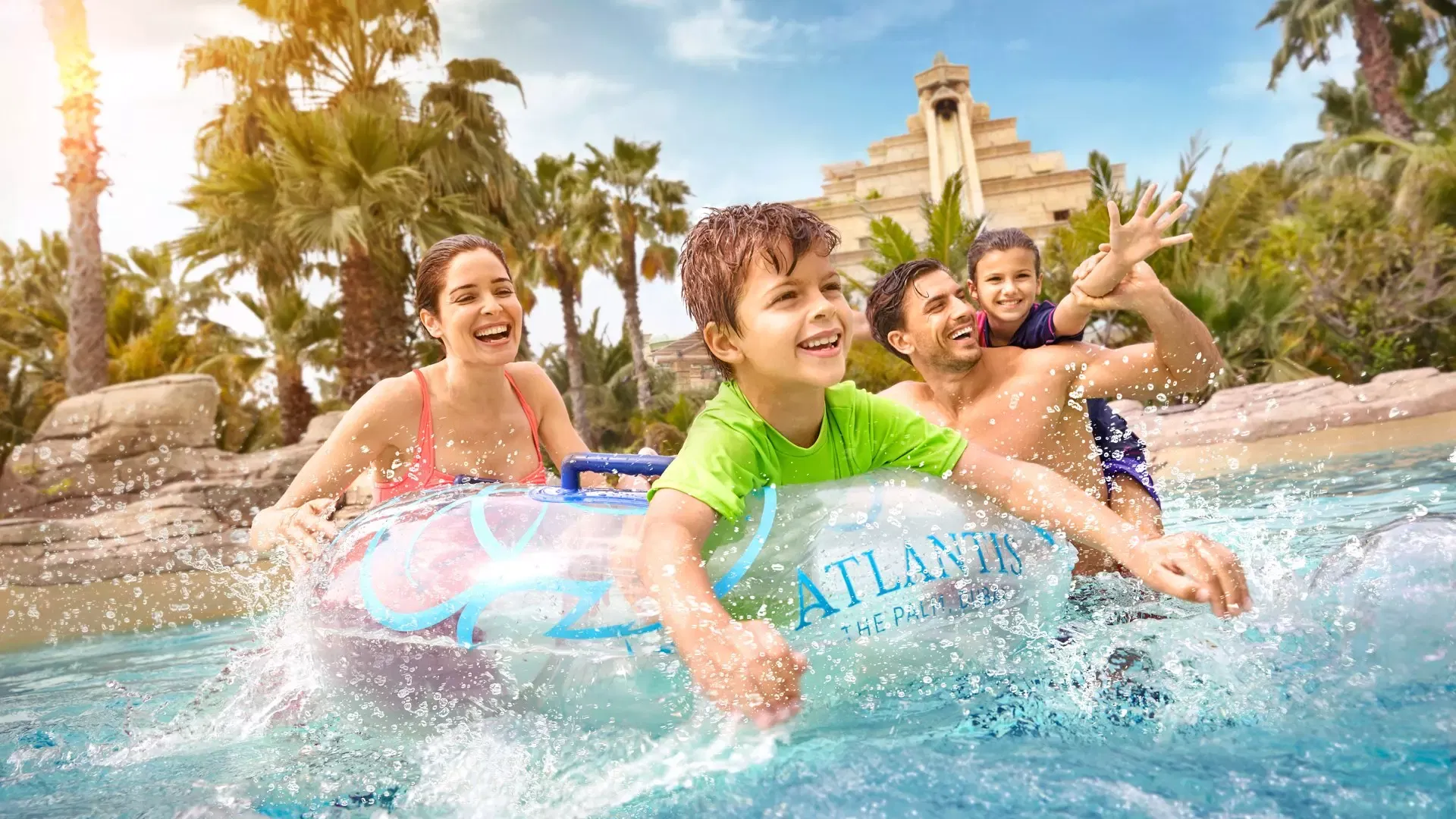 A smiling family of four riding an Atlantis-branded inflatable ring down the rapids past palm trees and a stone ziggurat tower at Aquaventure Waterpark