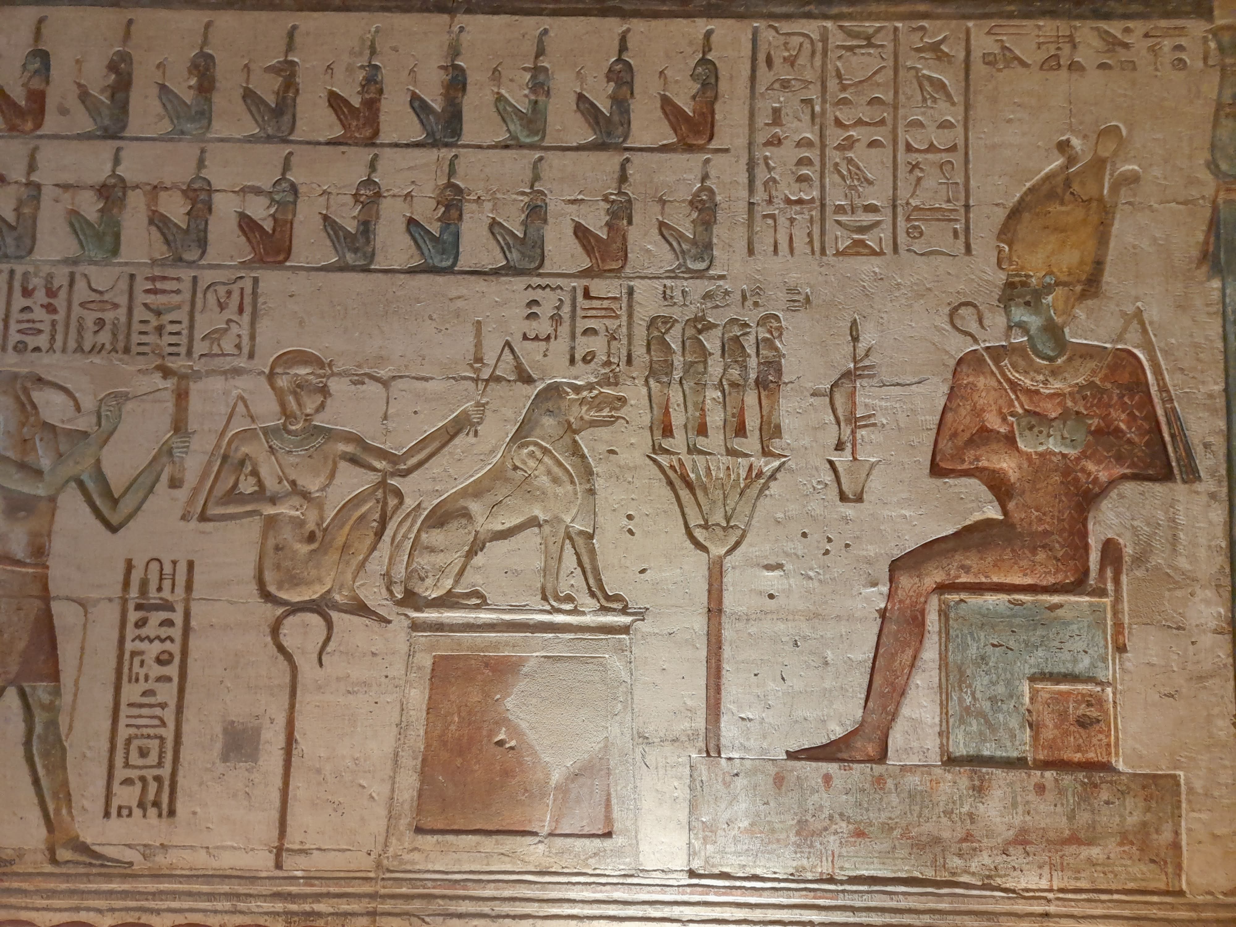 Detailed ancient wall carving showing a seated pharaoh and deities before offering tables in reddish-brown paint